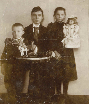 [Translate to Niederländisch:] Schwarz-Weiß-Aufnahme. Grete im dunklen Kleid und mit Puppe im Arm neben ihren Brüdern. Curt im Anzug, Johannes als kleiner Junge im Ringelpullover.