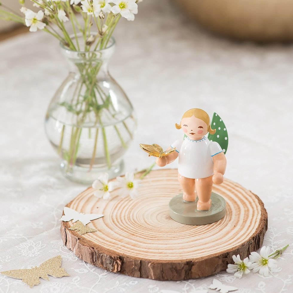 Elfpunkte-Engel mit Schmetterling auf einer Holzscheibe. Daneben ein kleiner Strauß weißer Blumen