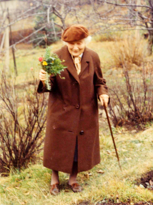 [Translate to Niederländisch:] Grete Wendt im braunen Mantel und mit Mütze mit einem bunten Blumenstrauß in der Hand. Sie steht in einem sonst herbstlichen Garten.